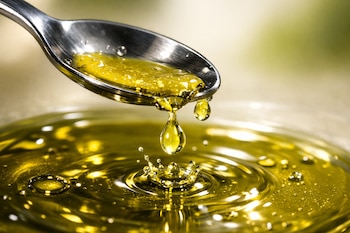 Cuchara metálica vertiendo jugo de limón amarillo brillante sobre aceite de oliva virgen extra, creando una salpicadura con pequeñas gotas en una toma macro de cerca.
