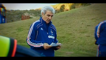 Un hombre de cabello gris con chándal azul de la selección francesa sostiene papeles al aire libre, con una colina verde de fondo