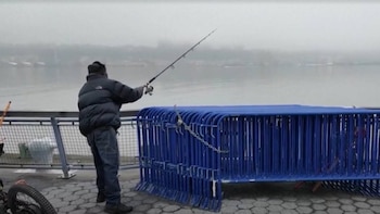 Hombre de espaldas pescando en un muelle. Vallas metálicas azules apiladas a la derecha y un cuerpo de agua brumoso al fondo