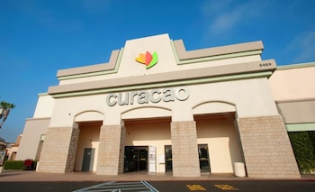 Vista exterior frontal de una tienda Curacao de color beige con letras grises y un logo colorido, bajo un cielo azul brillante