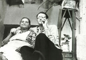 Fotografía en blanco y negro de un hombre joven con gafas y una mujer mayor sentados, sonriendo, con plantas en macetas al fondo