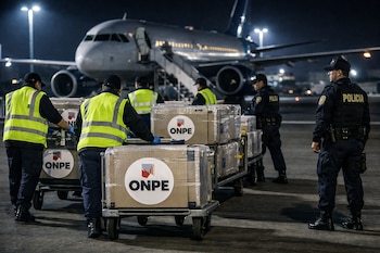 Vista nocturna en un aeropuerto donde personal con chalecos amarillos y policías uniformados mueven cajas con el logo de ONPE, con un avión de fondo.