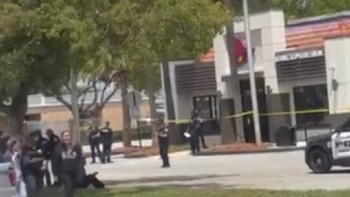Una escena con oficiales de policía, un coche patrulla, cinta amarilla y varias personas en una calle con un edificio, posiblemente un Taco Bell, y árboles