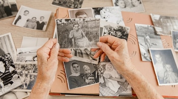 Primer plano de manos sosteniendo una foto en blanco y negro de una mujer en un campo, con otras fotos y un álbum de fotos dispersos sobre una mesa de madera.
