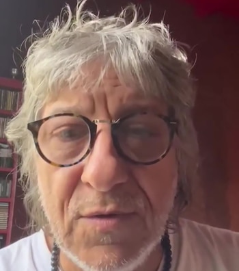 Primer plano de un hombre senior con cabello gris desordenado y barba incipiente, usando gafas de carey y un collar de cuentas oscuras