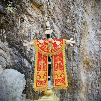 Cruz negra con detalles plateados y acrónimo INRI, adornada con tela roja y dorada que muestra bordados florales, cruces y cálices, sobre fondo rocoso con una roca y vegetación