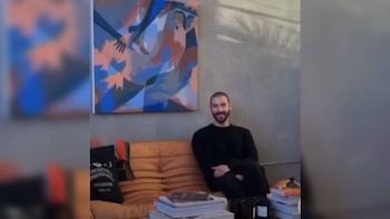 Un hombre barbudo con camiseta negra sentado en un sofá marrón, sonriendo a la cámara, con un cuadro abstracto colorido en la pared