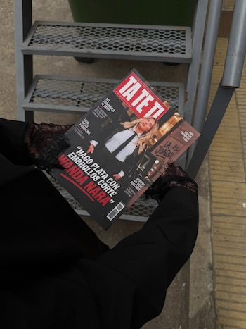 Unas manos con mangas de encaje negro sostienen una revista con la fotografía de una mujer rubia y varios titulares en la portada