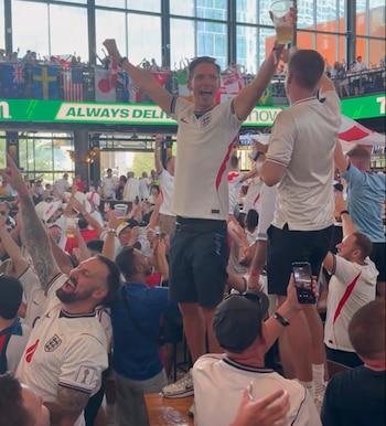 Un grupo de hombres con camisetas de Inglaterra celebra con los brazos en alto en un bar, algunos de pie sobre las mesas, con banderas internacionales de fondo