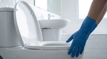 Primer plano de dos manos con guantes de goma azules extendiéndose hacia un inodoro blanco con la tapa abierta en un baño.
