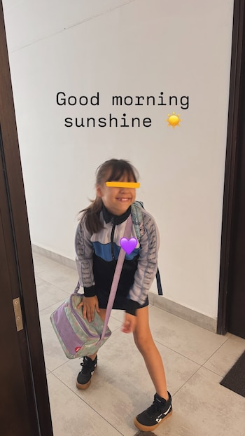 Una niña en uniforme escolar sostiene una lonchera frente a una pared blanca, con el texto en inglés "Good morning sunshine" en la parte superior