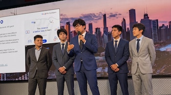 Cinco hombres jóvenes en traje sobre un escenario; uno habla con micrófono frente a una pantalla con texto y el logo de Google for Developers.