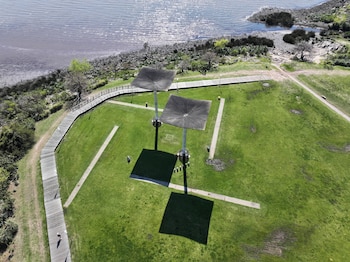 Vista aérea de un parque de césped verde con dos estructuras metálicas que proyectan sombras cuadradas, pasarelas de madera y la orilla de un río