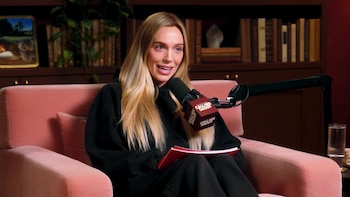 Mujer rubia sentada en sillón rosa, con sudadera negra, micrófono Call Her Daddy, cuaderno rojo. Fondo con estanterías y un vaso de agua