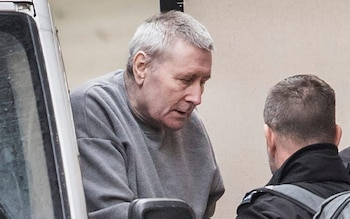 Un hombre de mediana edad con pelo gris y sudadera gris mira hacia abajo, junto a un vehículo blanco y la espalda de una persona con uniforme oscuro