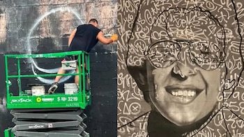 Un hombre pinta un muro negro sobre una plataforma de tijera verde junto a un mural en blanco y negro con el rostro de una persona con gafas