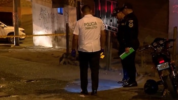Tres hombres, uno con camiseta de criminalística y dos con uniforme policial oscuro, realizan pericias en una calle de noche con cinta policial, un coche y una motocicleta
