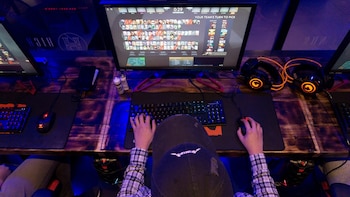 Vista superior de una persona con gorra que manipula un teclado y un ratón frente a un monitor de computadora en una mesa de madera