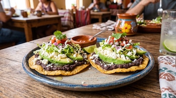 Dos tostadas con frijoles, aguacate, lechuga y queso fresco en un plato, junto a salsa roja y una rodaja de lima sobre una mesa de madera.