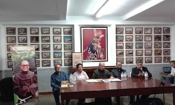 Hombres sentados ante una mesa con botellas de agua, un cartel vertical y paredes cubiertas con cuadros enmarcados