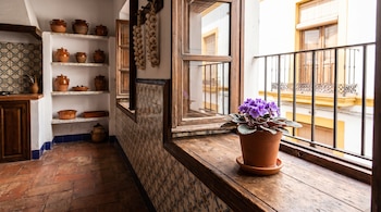 Una maceta con una violeta africana morada sobre un alféizar de madera junto a azulejos, estantes con vasijas de barro y ristras de ajos.