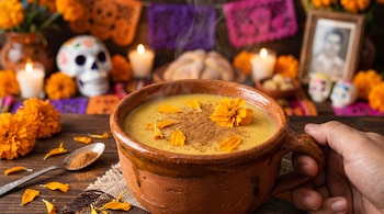 Taza de barro con bebida amarilla decorada con canela y pétalos, sostenida por una mano frente a velas, calaveras de azúcar y flores.