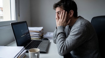 Hombre con suéter gris, manos en la cara y expresión de preocupación, frente a un laptop que muestra 'Actualizando... 12%', en un escritorio con papeles y una taza.