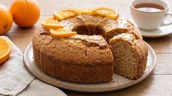 Un budín de naranja con glaseado y rodajas de naranja cortada sobre un plato, con una porción separada. Alrededor hay naranjas y una taza de té.