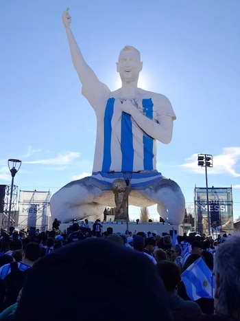 Vista de una enorme estatua de Lionel Messi vestido con la camiseta argentina, con el brazo levantado, frente a una multitud de personas con banderas