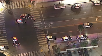 Vista aérea nocturna de una intersección con varias patrullas de policía con luces intermitentes, personas y cinta de precaución en la calle
