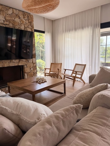 Un salón con chimenea de piedra y televisión, sofá beige con cojines, mesa de centro de madera, dos sillas de mimbre y grandes ventanales con cortinas transparentes