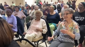 Un grupo de adultos mayores sentados en sillas participan en una actividad dentro de un salón iluminado