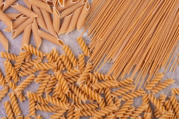 Vista superior de pasta integral seca. Se observan fideos penne, fusilli y espagueti de color marrón claro sobre un fondo gris