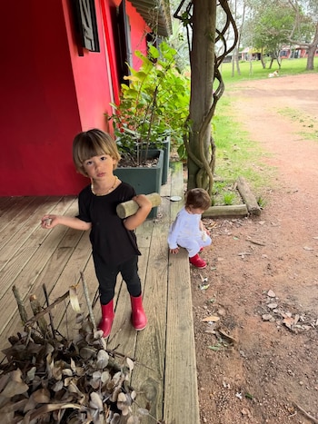 Dos niños en una terraza de madera junto a una pared roja. Uno, con camiseta negra y botas rojas, sostiene un trozo de madera; el otro, de blanco y botas rojas, se agacha