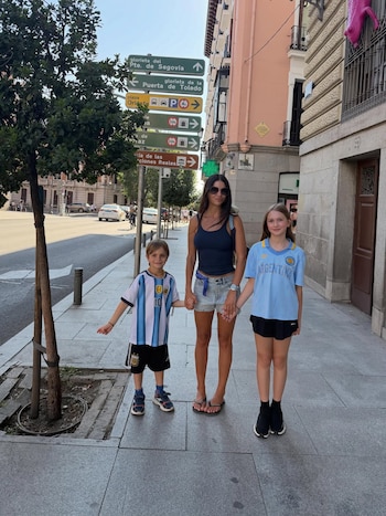 Zaira Nara y sus dos hijos, una niña y un niño, caminan por una acera. Se ven señales de tráfico, un árbol, edificios y vehículos en la calle