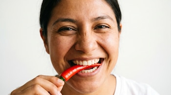 Primer plano de una persona mexicana sonriente con pelo oscuro, mordiendo un chile rojo brillante que sostiene con su mano sobre un fondo claro.