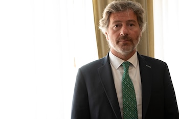 Hombre maduro con pelo canoso y barba, vistiendo un traje oscuro, camisa blanca y corbata verde. Fondo brillante con cortina beige