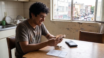 Joven con cabello rizado cuenta billetes sentado en una mesa de madera. Hay monedas, una cartera, un documento y una cocina detrás. Ventana con vista a la calle.