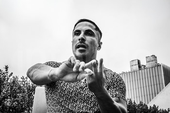 Un hombre, Ariel Fefer, sentado y gesticulando con ambas manos. Viste una camisa estampada. Al fondo, vegetación, un edificio y cielo claro. Fotografía en blanco y negro