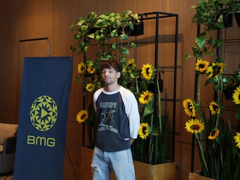 Un hombre de cabello oscuro y barba corta, vestido con camiseta bicolor y jeans, de pie frente a un fondo con girasoles, plantas y un banner de BMG