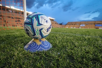 Balón de fútbol blanco, azul y verde sobre soporte amarillo y azul en campo de césped. Red de arco y tribunas iluminadas al fondo