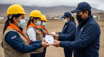 Cuatro personas con mascarillas, cascos amarillos, chalecos y chaquetas azules, intercambian documentos en un campo abierto con montañas peruanas al fondo.