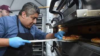 Un chef con camisa azul y delantal negro, usando guantes azules, introduce piezas de masa redondas cubiertas de sésamo en un horno industrial con una pala metálica