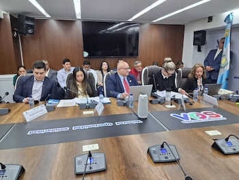 Grupo de ejecutivos y delegados en una mesa de reuniones grande, con laptops, micrófonos y una pantalla de televisión oscura al fondo de la sala