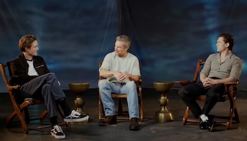Tres hombres, Robert Pattinson, Matt Damon y Tom Holland, están sentados en sillas de madera en un escenario con un fondo azul oscuro y luces sutiles
