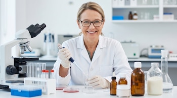 Una científica sonriente de mediana edad con gafas y bata blanca manipula una pipeta y un vaso de precipitados en un laboratorio lleno de instrumentos y muestras.