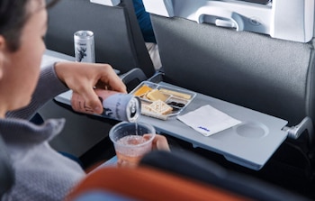Manos de una persona vertiendo bebida de una lata en un vaso plástico, una bandeja de asiento de avión con aperitivos y una servilleta, y otra lata