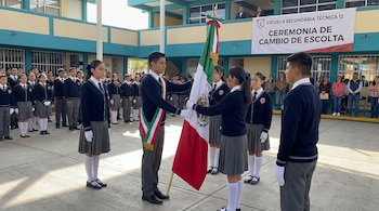 Jóvenes estudiantes con uniforme escolar realizan una ceremonia cívica en un patio, un alumno entrega una bandera mexicana a otro estudiante, varios alumnos observan.