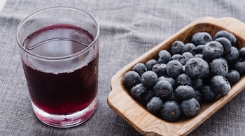 Un vaso de cristal con líquido rojizo oscuro y condensación, junto a un cuenco de madera con bayas de color azul oscuro sobre una tela gris.