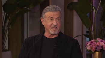 Sylvester Stallone viste ropa oscura y habla frente a un fondo con plantas de interior y un arreglo floral rosado